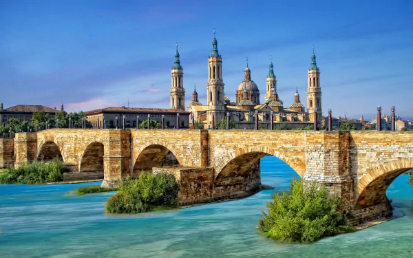 HD PC desktop background: Zaragoza, Spain cityscape with a man-made stone bridge over a turquoise river and historic basilica towers beneath a clear blue sky.