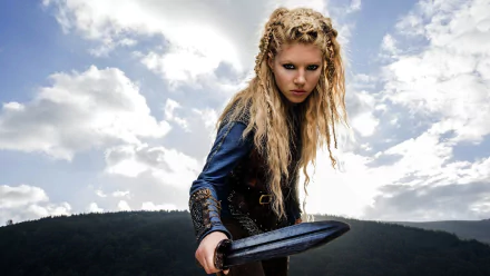 HD PC desktop wallpaper: blonde braided shieldmaiden from the TV show Vikings, fierce gaze as she holds a sword against a cloudy sky and distant hills.