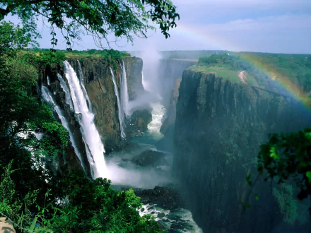 HD desktop wallpaper of Victoria Falls showcasing roaring waterfalls cascading down green cliffs surrounded by lush forest and a vibrant rainbow arching over the scene.