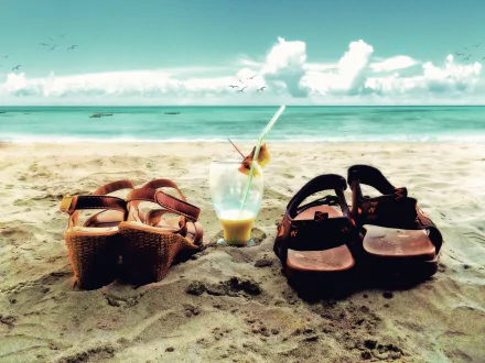 Two pairs of sandals next to a refreshing glass drink on the beach, with a serene horizon and romantic clouds in the background. A tranquil holiday vibe captured in this HD wallpaper.