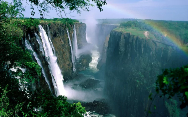 HD desktop wallpaper of Victoria Falls showcasing roaring waterfalls cascading down green cliffs surrounded by lush forest and a vibrant rainbow arching over the scene.