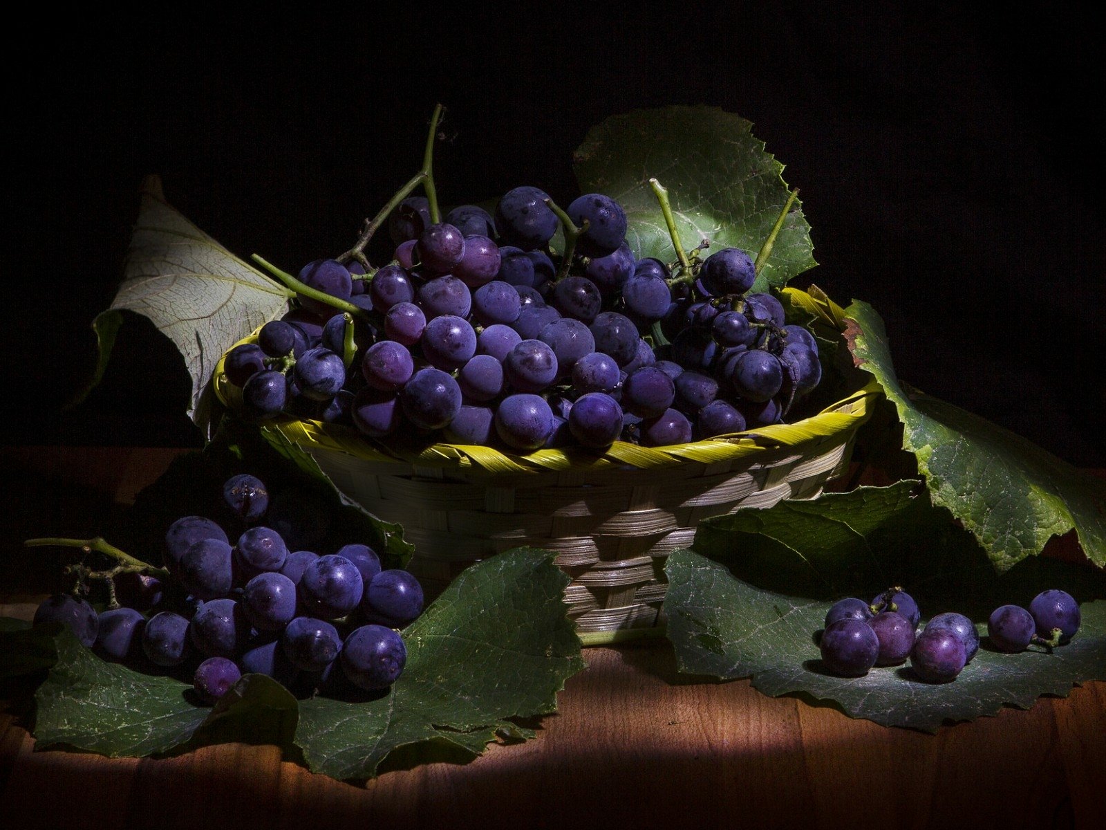A still life composition featuring a wicker basket filled with dark purple grapes, surrounded by vibrant green leaves, set against a dark background, capturing rich textures and colors.