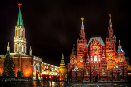 Night view of illuminated historic buildings in Moscow's Red Square, showcasing iconic Russian architecture and church structures against a dark sky.
