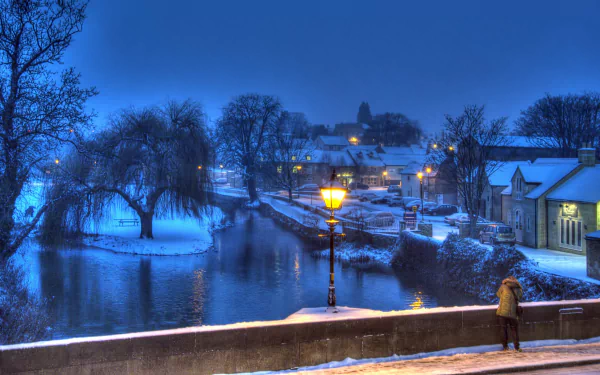 4K Ultra HD winter night scene of a quiet town with a softly lit bridge over calm water, snow-covered trees, and warmly glowing streetlights.