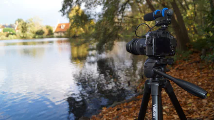 A Sony camera on a tripod, equipped with a microphone, overlooks a serene pond surrounded by trees. The image captures the tranquil beauty of the lake setting in stunning 4K Ultra HD.
