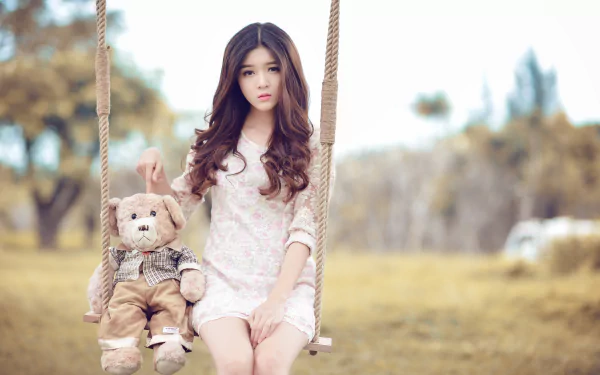 A brunette Asian woman is seated on an outdoor swing next to a teddy bear. The background features bokeh effects, providing a serene and dreamy atmosphere. This HD image serves as a desktop wallpaper.