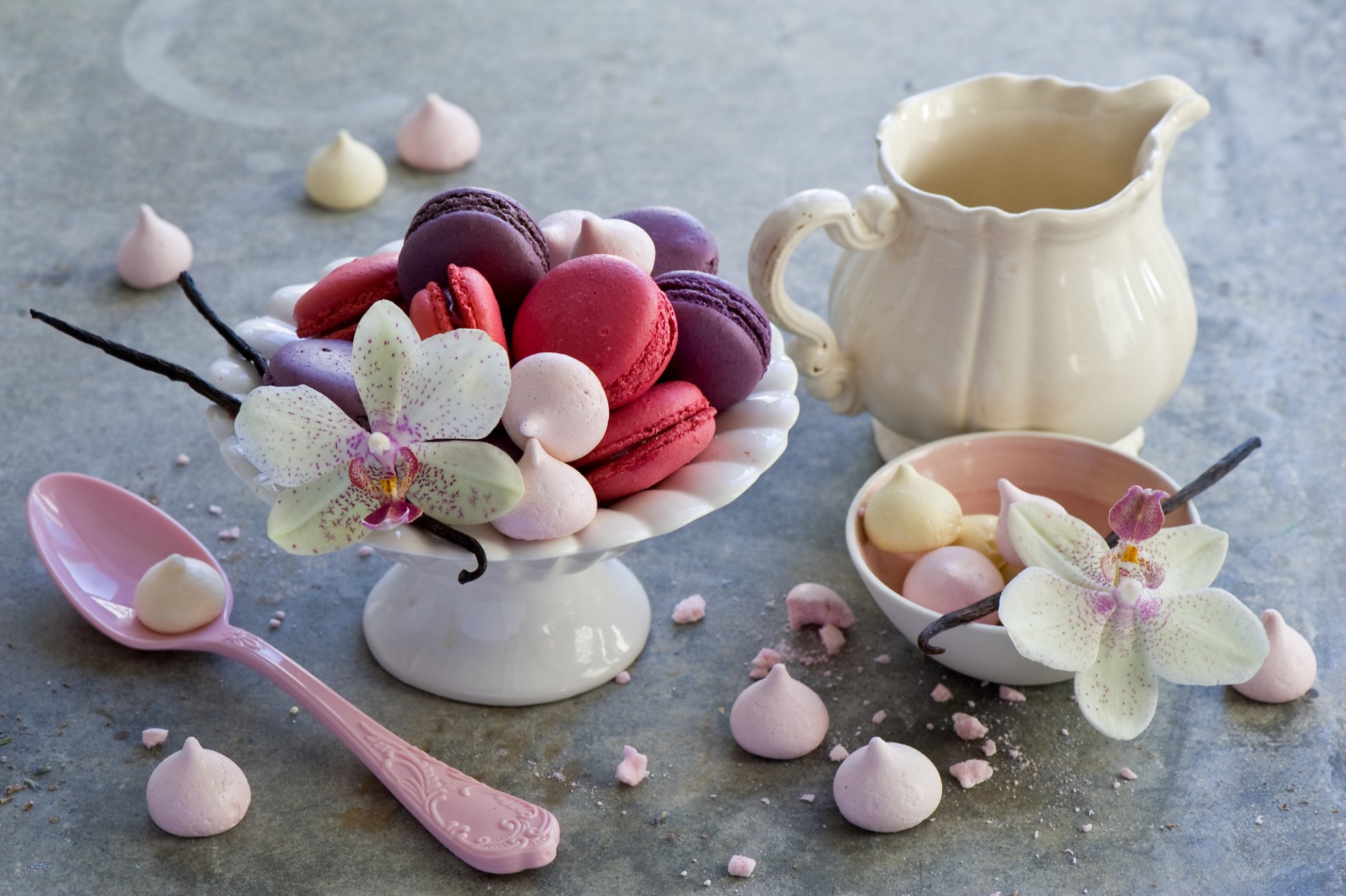 A beautifully arranged still life of colorful macarons in a decorative bowl, accompanied by delicate meringues and an elegant pitcher, set against a soft, textured background.