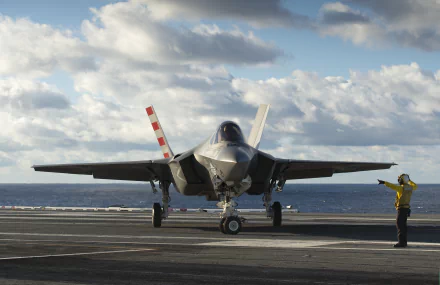 Lockheed Martin F-35 Lightning II jet fighter on an aircraft carrier deck with a crew member signaling, set against a cloudy sky and ocean backdrop.