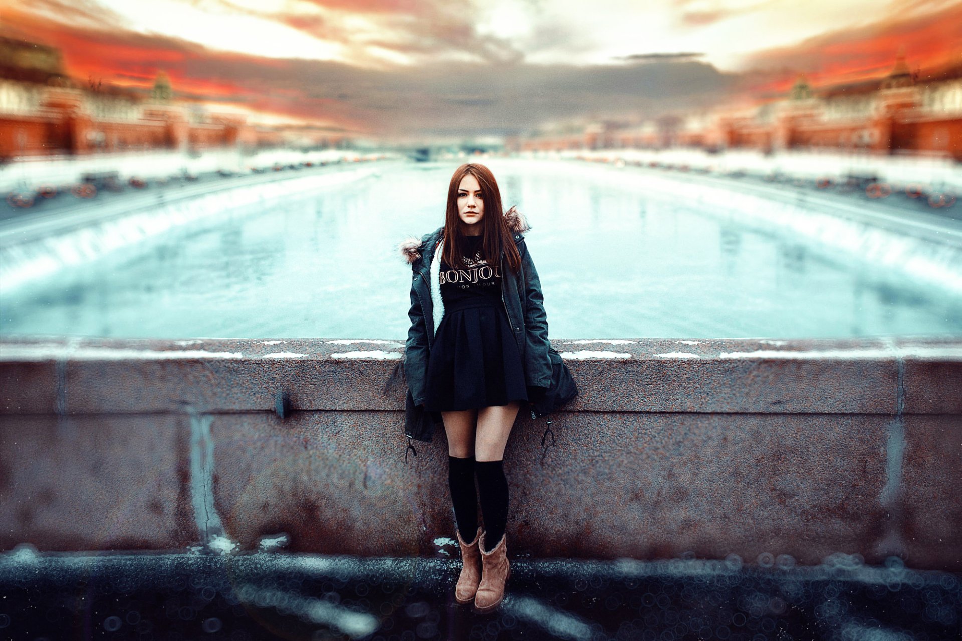 HD desktop wallpaper featuring a brunette woman model standing outdoors by a blurred waterfront with vibrant colors and a dramatic sky.