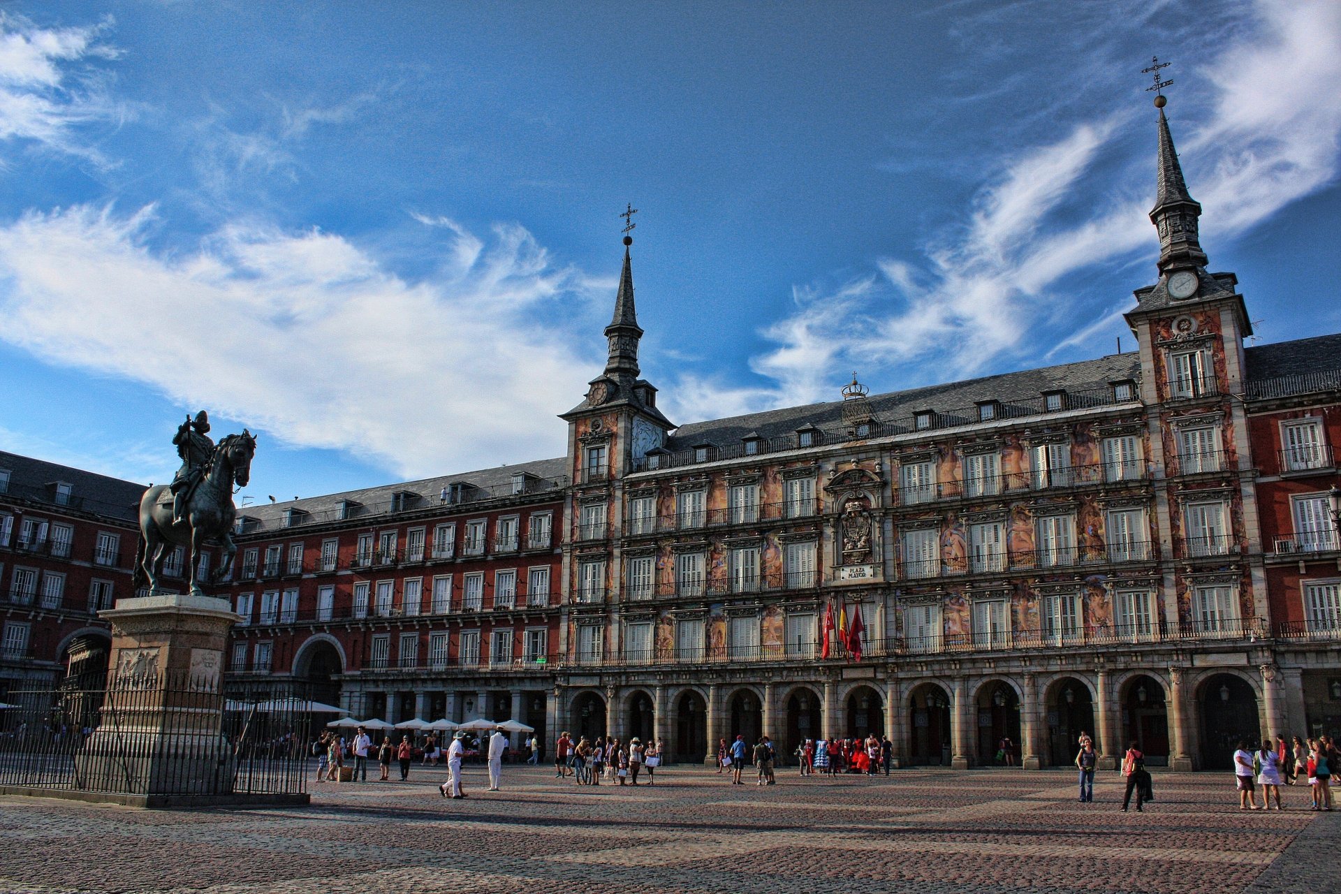 A 4K Ultra HD image of a historic square in Madrid, Spain, featuring a man-made statue of a man on horseback and a grand building under a vibrant blue sky.