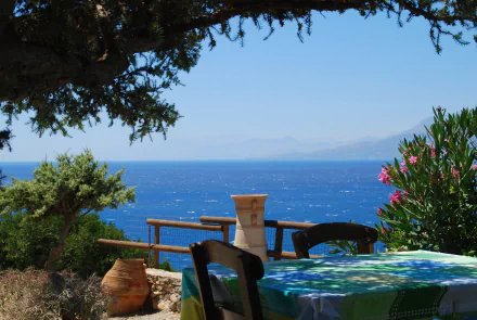 Scenic view of a table overlooking the blue ocean in Crete, framed by trees and flowering plants, captured in HD photography as a desktop wallpaper background.