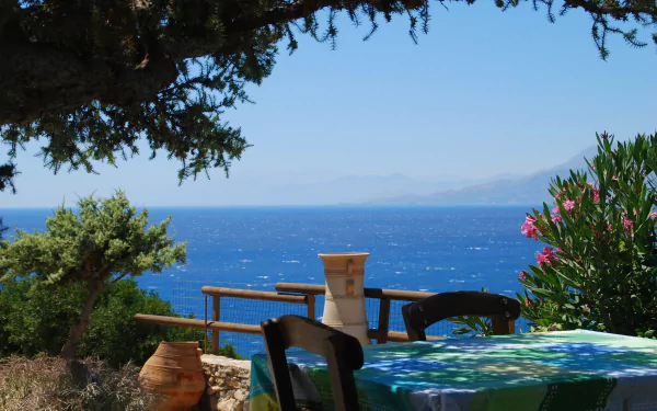 Scenic view of a table overlooking the blue ocean in Crete, framed by trees and flowering plants, captured in HD photography as a desktop wallpaper background.