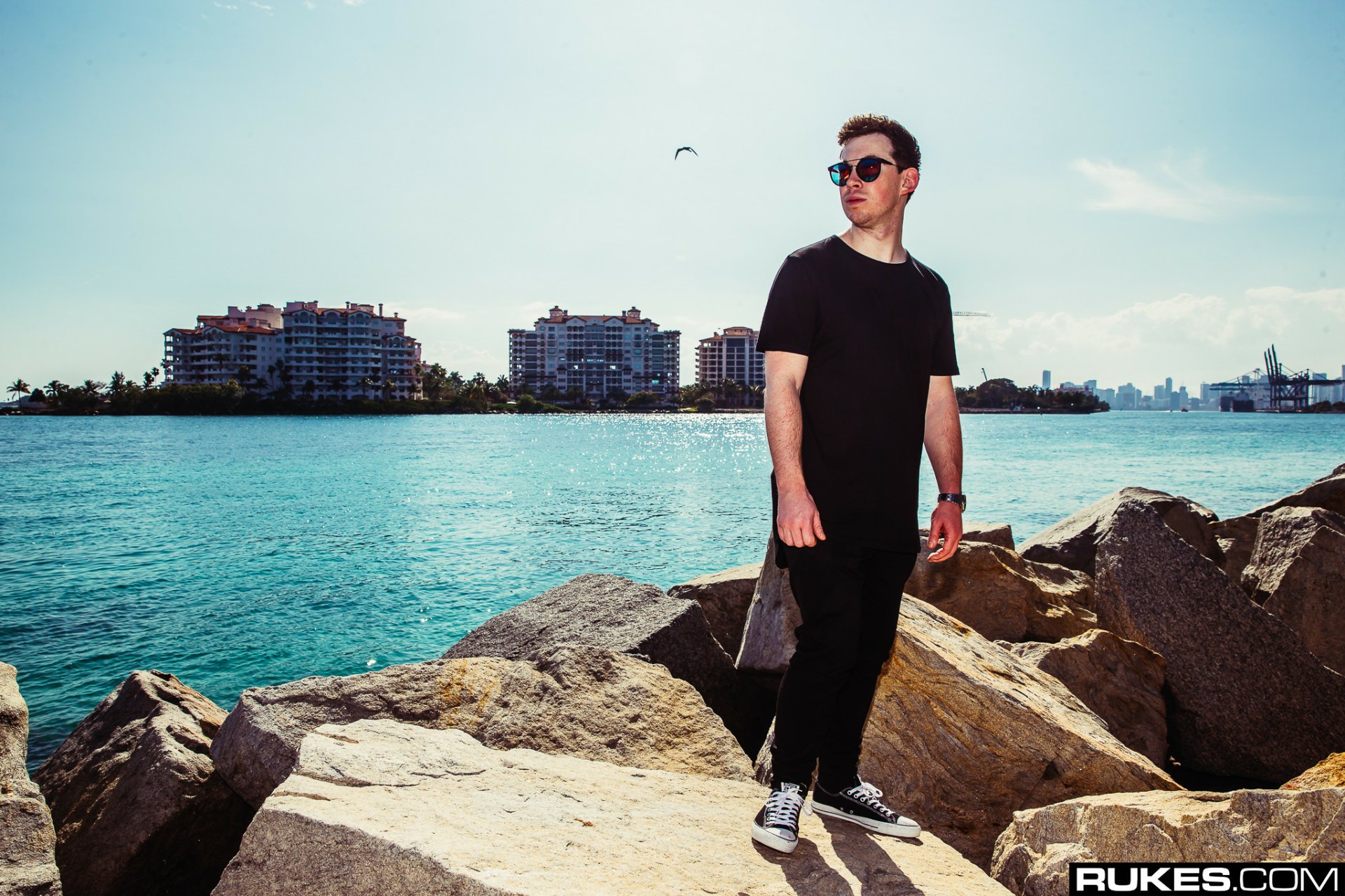HD PC desktop wallpaper of a DJ in black and sunglasses standing on sunlit coastal rocks with turquoise water and a city skyline behind, projecting energetic electronic music vibes.