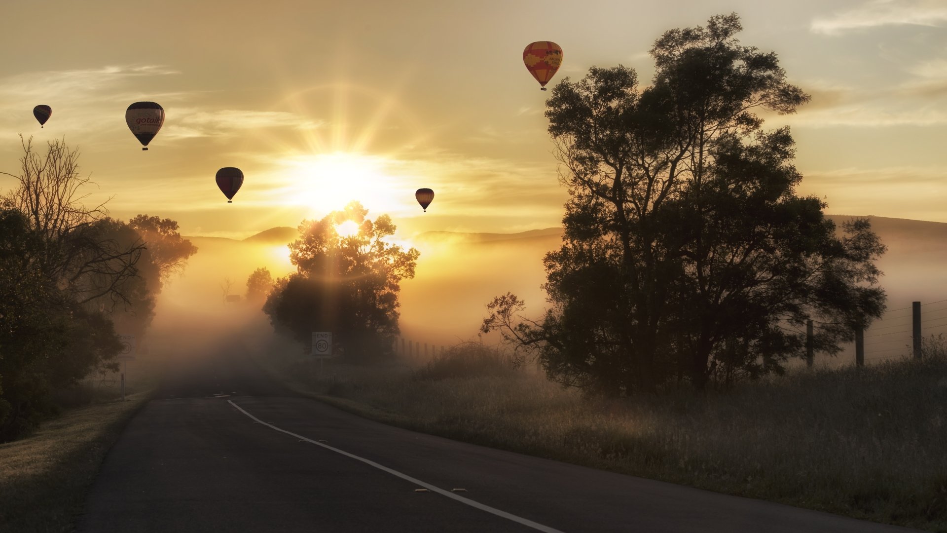 A serene landscape at sunrise, featuring a winding road through fog, silhouetted trees, and colorful hot air balloons rising against the warm sunbeams. 4K Ultra HD wallpaper.