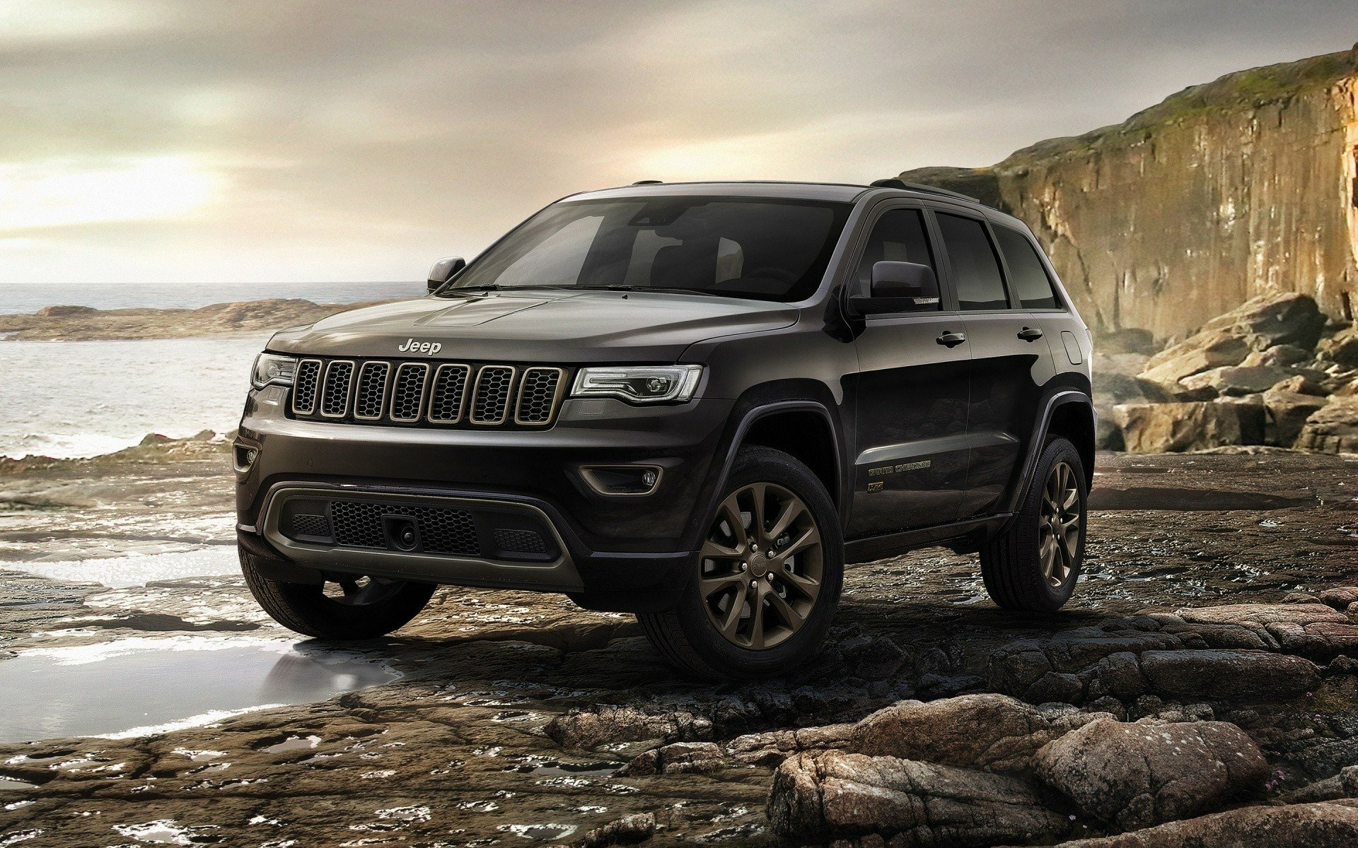 Black Jeep Cherokee SUV on a rocky shoreline with cliffs and ocean at dusk — HD PC desktop wallpaper and background.