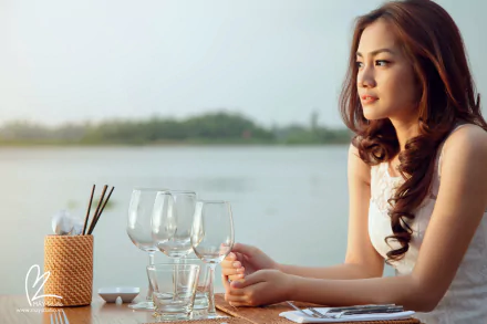HD desktop wallpaper featuring an Asian woman gazing thoughtfully over a serene lake, with empty glasses and a wicker container with utensils on a wooden table in the foreground.