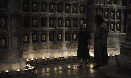 Arya Stark and Jaqen H'ghar stand in a dimly lit room adorned with faces, captured in a striking scene from Game of Thrones. This HD wallpaper brings their connection to life.
