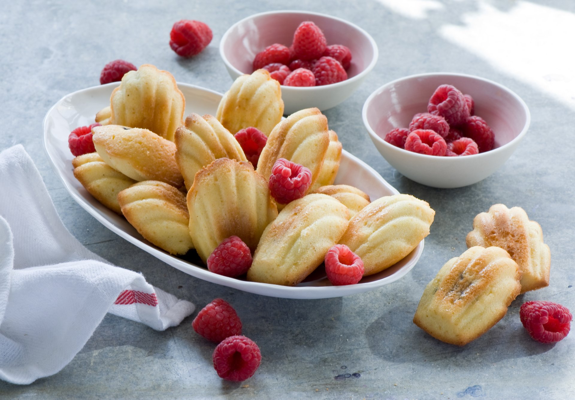 A 4K Ultra HD still life of golden Madeleines and fresh raspberries arranged in a white dish and bowls on a gray surface, creating a vibrant food display.