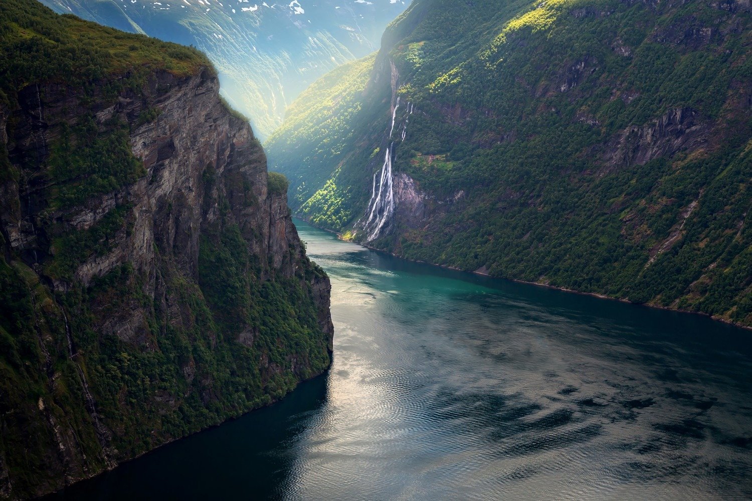 HD PC desktop wallpaper: aerial view of a Norwegian fjord — a turquoise lake winding between steep mossy mountains, with a distant waterfall and sunlight filtering into the valley.