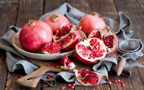 HD PC desktop wallpaper still life of pomegranate fruit and food: whole and halved pomegranates with jewel-red seeds on a wooden board and gray cloth, a small sieve beside them.