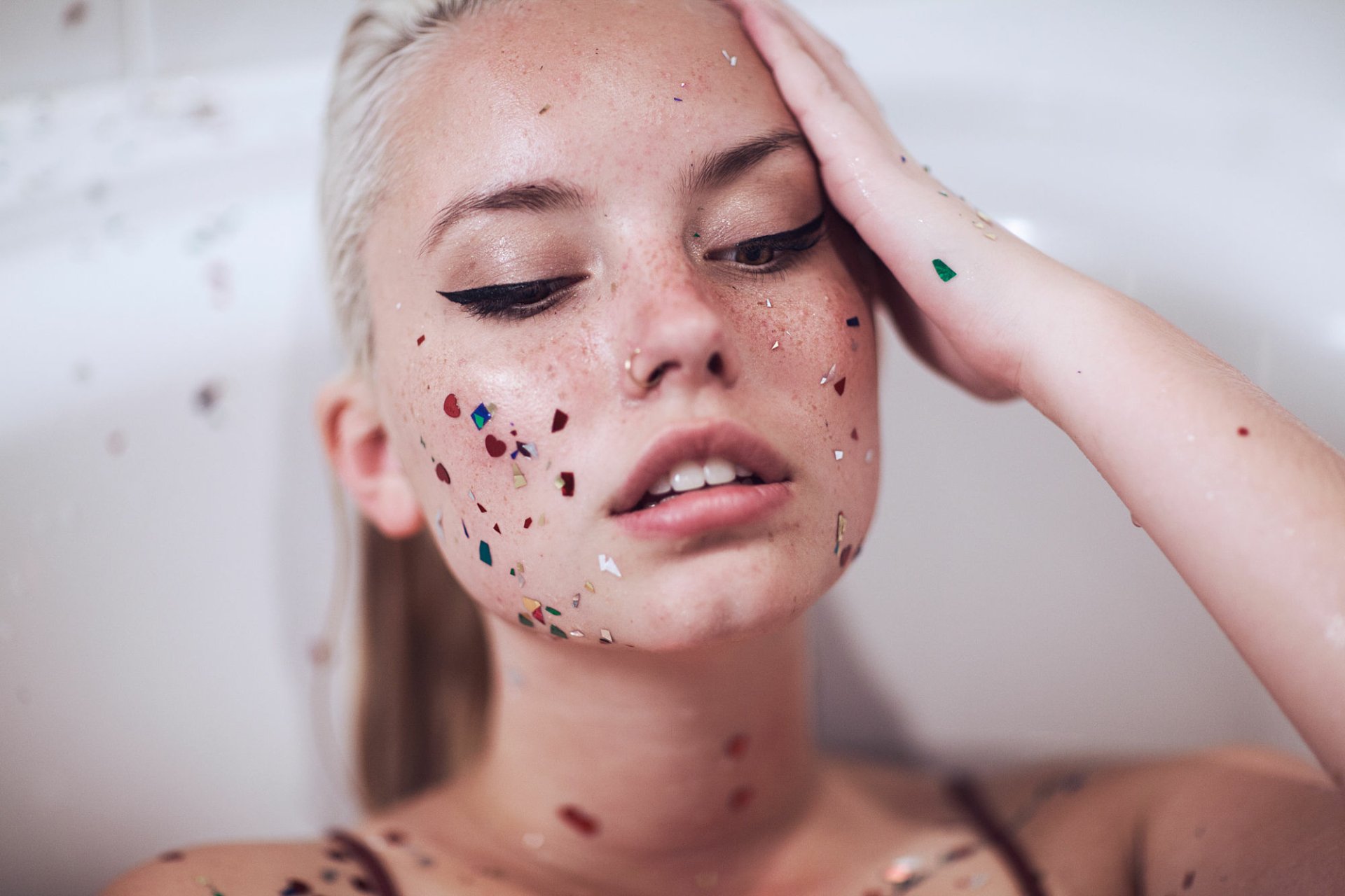 Close-up of a blonde woman with brown eyes and freckles, her face adorned with colorful confetti, captured in HD as a striking desktop wallpaper background.