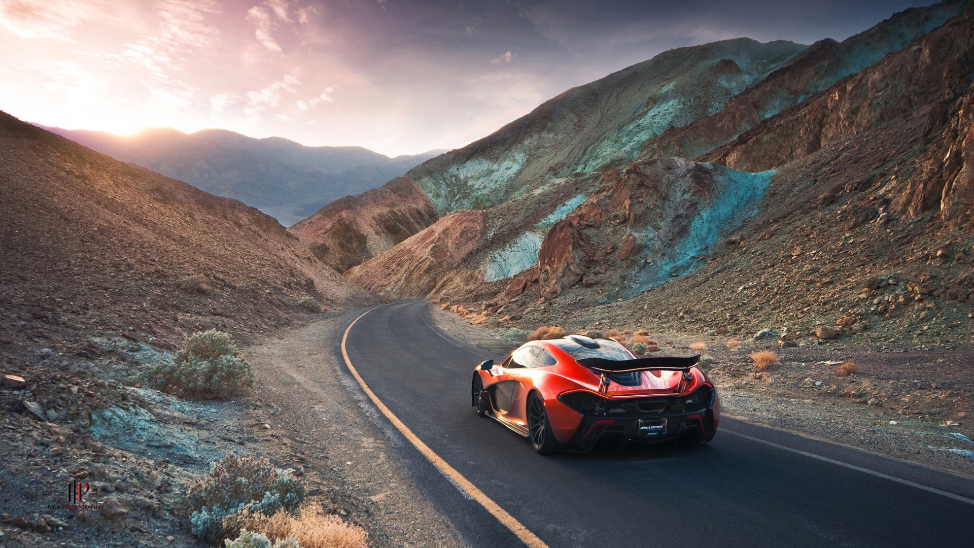 A stunning McLaren P1 supercar in vibrant orange, navigating a scenic winding road through mountainous terrain, with a breathtaking sunset illuminating the landscape.