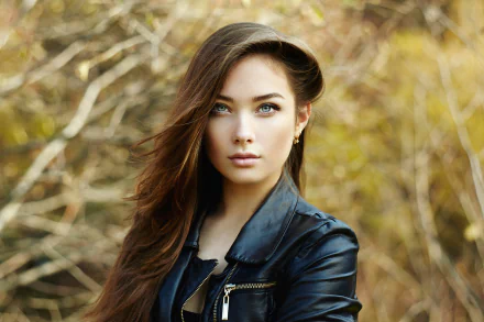 HD desktop wallpaper of a brunette woman model with striking blue eyes, wearing a black leather jacket, set against a softly blurred natural background.
