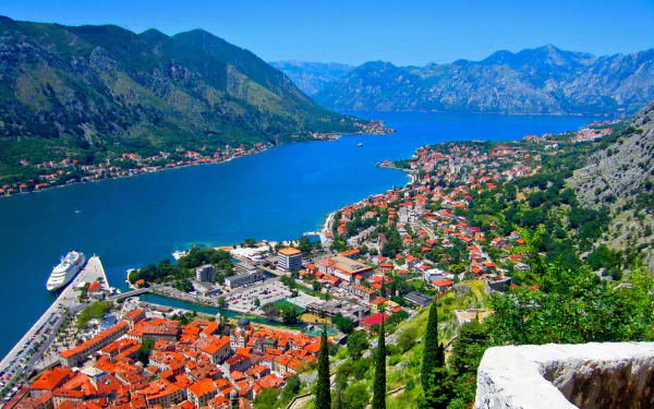 A panoramic coastal view of Kotor, Montenegro, showcasing the town's red-roofed houses nestled between mountains and the deep blue waters of the bay.