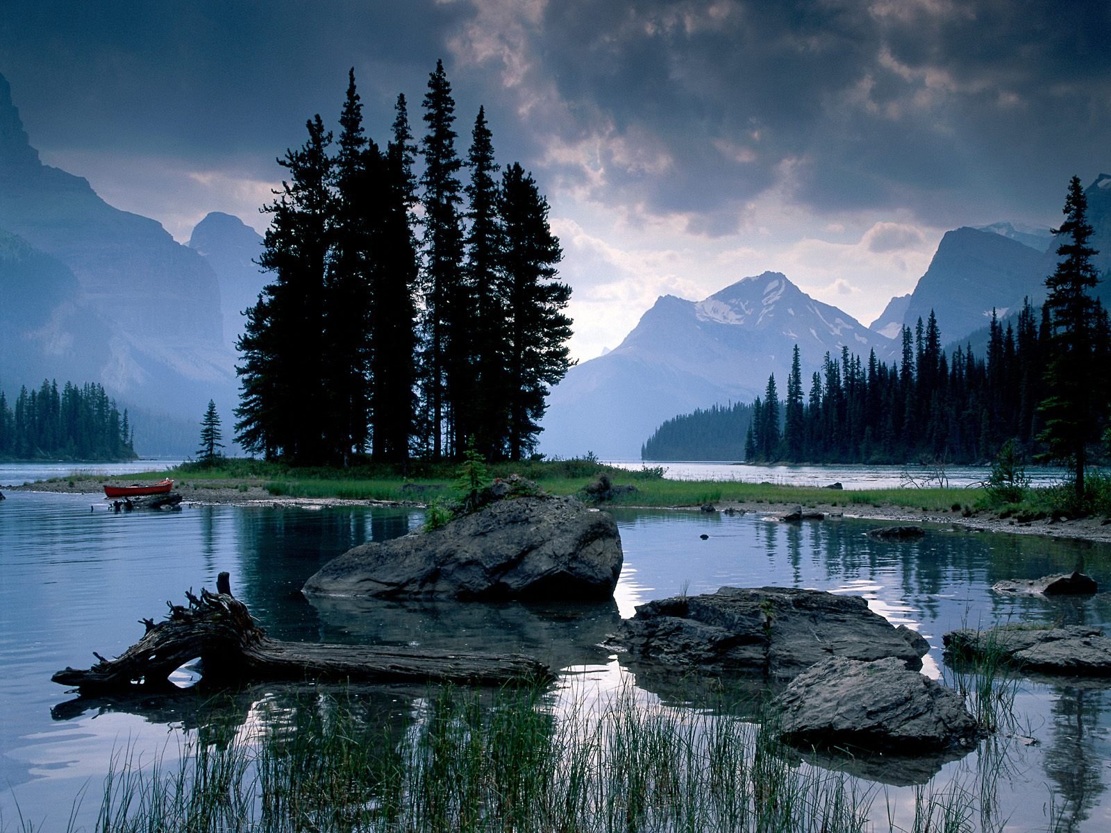 A serene HD desktop wallpaper showcasing a tranquil lake surrounded by lush trees and majestic mountains under a moody sky, capturing the essence of nature's beauty.