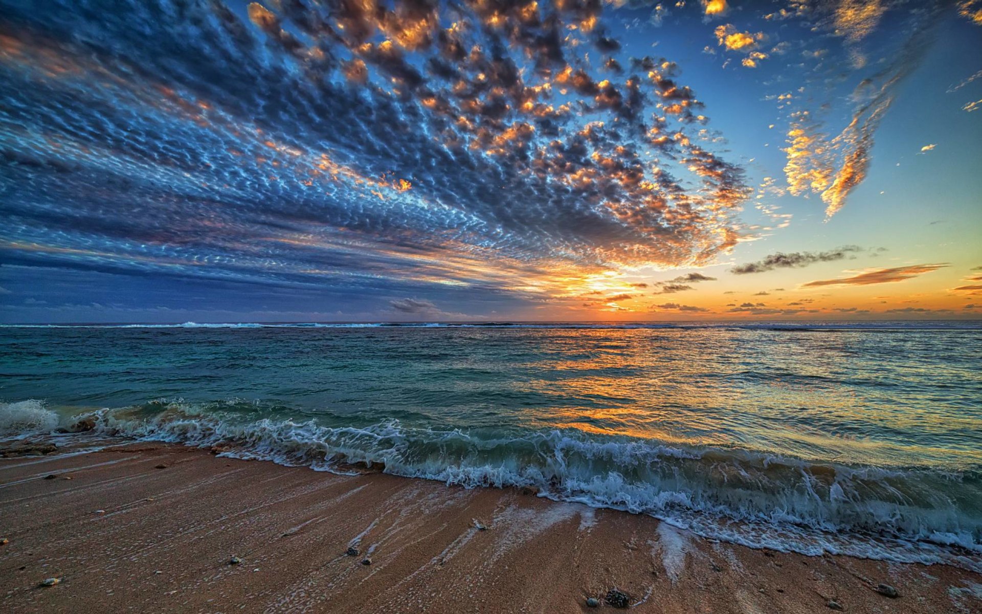 Vibrant HD desktop wallpaper of a beach at sunset, showcasing dramatic clouds over the ocean and the serene sky meeting the sea along the sandy shore.