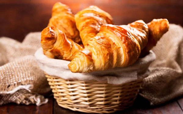 5K Ultra HD PC desktop wallpaper: golden flaky croissants in a wicker basket on rustic cloth, breakfast food close-up.