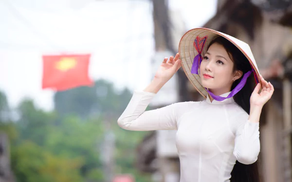 A Vietnamese woman wearing a traditional white Ao Dai and an Asian conical hat, captured in 4K Ultra HD against a blurred background with the Vietnamese flag.