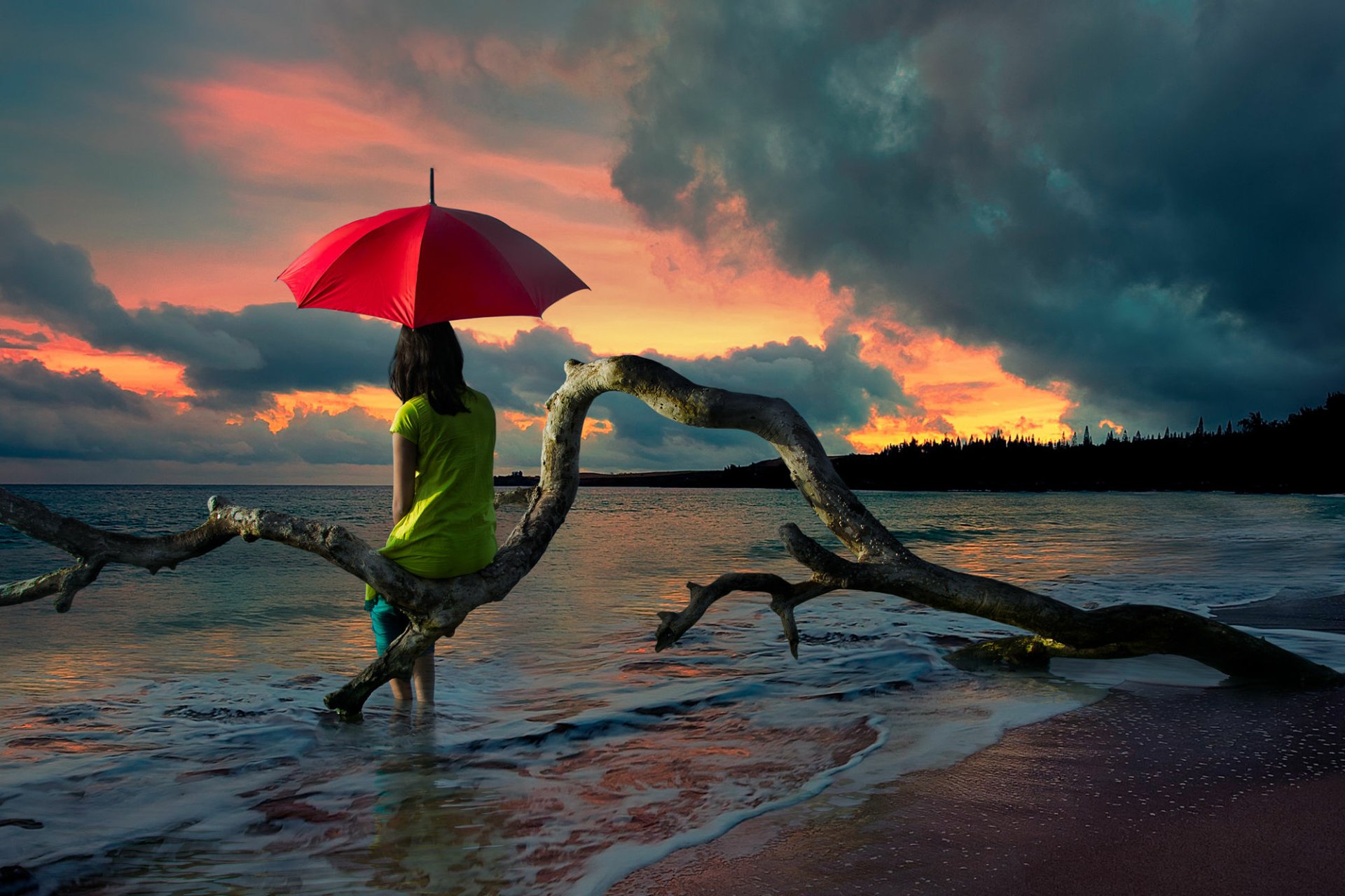 A woman holding a red umbrella stands at the shore during a stunning sunset, gazing at the ocean. A weathered branch arches over the water as waves gently lap the sand. HD desktop wallpaper background.