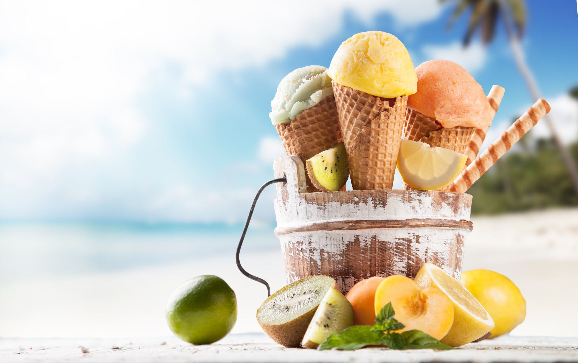 A vibrant assortment of ice cream cones in a basket, surrounded by fresh lemons and kiwi, evokes a refreshing summer vibe against a sunny beach backdrop.