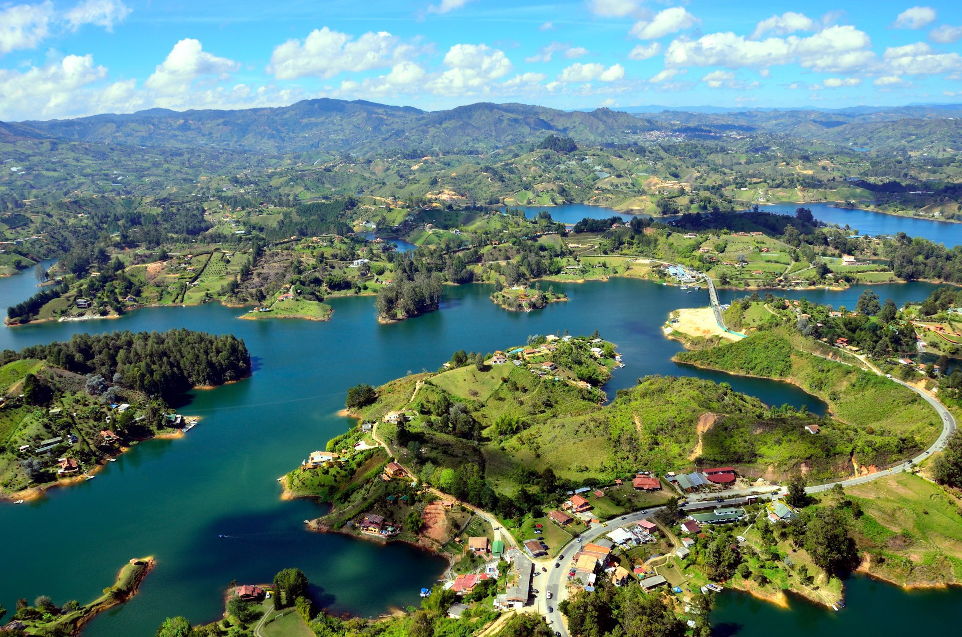 Aerial HD PC wallpaper showcasing a vibrant island village surrounded by water, lush green landscapes, and rolling hills under a bright, partly cloudy sky.