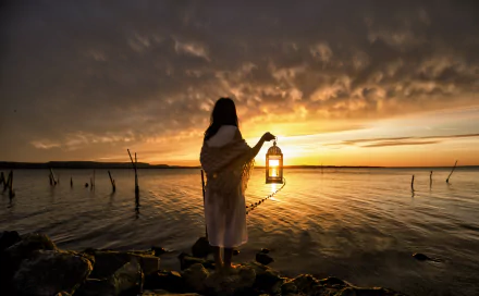 A woman stands at a lakeside during sunset, holding a lantern. The scene exudes tranquility and beauty, with golden tones reflecting on the water. This HD image makes a captivating desktop wallpaper and background.