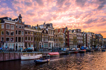 HD desktop wallpaper of a vibrant Amsterdam canal at sunset, featuring classic Dutch houses, moored boats, and a colorful sky in the Netherlands.