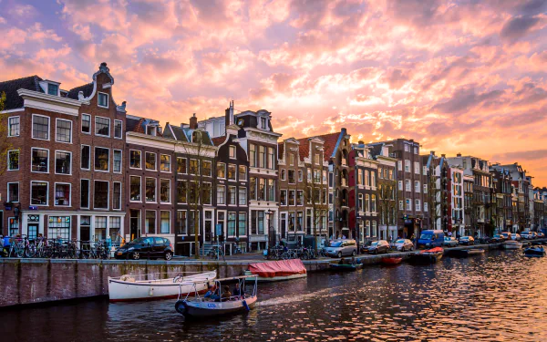 HD desktop wallpaper of a vibrant Amsterdam canal at sunset, featuring classic Dutch houses, moored boats, and a colorful sky in the Netherlands.