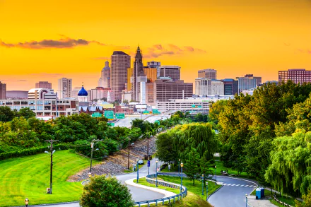 4K Ultra HD view of Hartford, Connecticut skyline at sunset with city buildings, a winding road, and lush trees in the foreground.