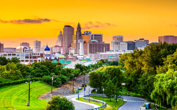 4K Ultra HD view of Hartford, Connecticut skyline at sunset with city buildings, a winding road, and lush trees in the foreground.