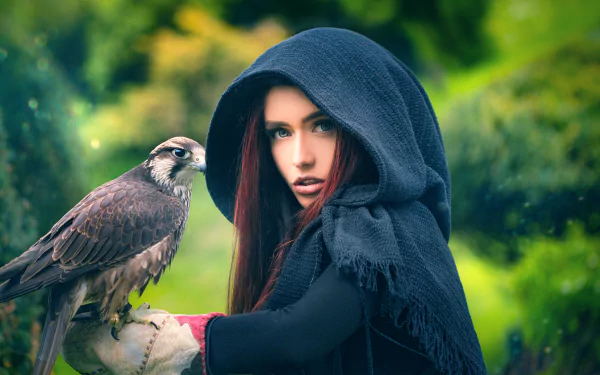 A red-haired woman model in a dark hood holds a hawk, set against a softly blurred green background, captured in high-definition for PC desktop wallpaper.
