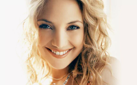 Close-up of American actress Kate Hudson with blonde hair, green eyes, and a radiant smile, set against a bright background in this HD desktop wallpaper.