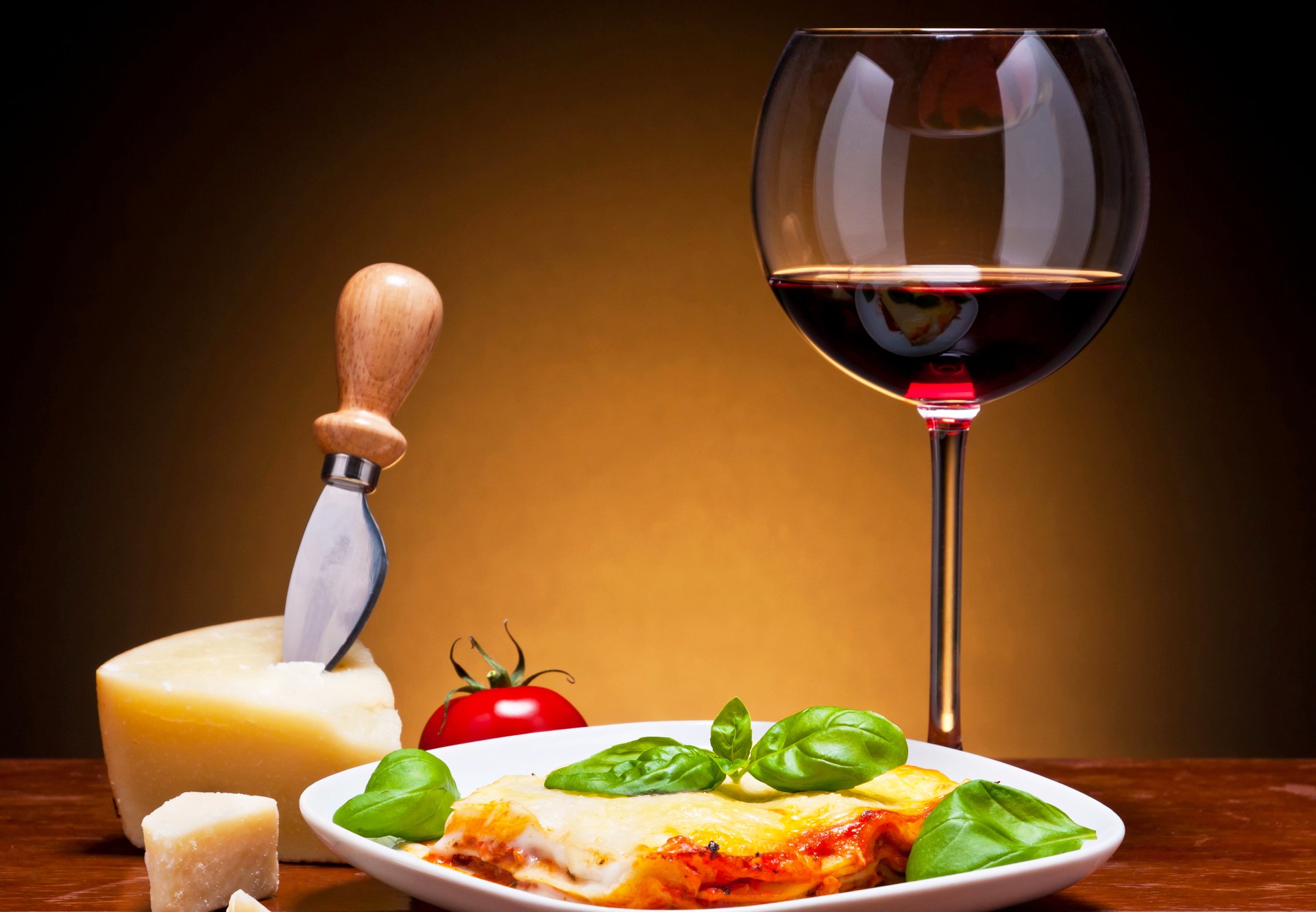 Cheesy lasagna with basil and tomato, a cheese wedge and knife, and a glass of red wine on a wooden table — 2K Quad HD PC desktop wallpaper and background.