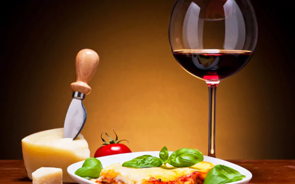 Cheesy lasagna with basil and tomato, a cheese wedge and knife, and a glass of red wine on a wooden table — 2K Quad HD PC desktop wallpaper and background.