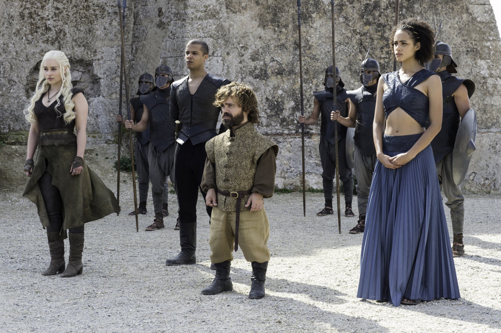 A scene from Game of Thrones featuring Daenerys Targaryen, Tyrion Lannister, Grey Worm, and Missandei, surrounded by guards, set against a stone backdrop.