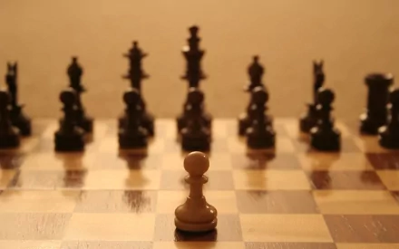 A close-up of a single white pawn in the foreground, surrounded by dark chess pieces, set against a warm, blurred background. This image serves as an elegant HD desktop wallpaper.