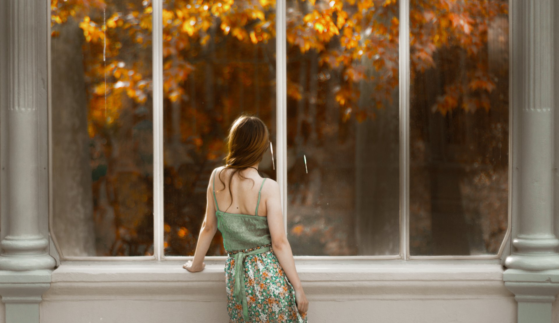 A redhead woman stands by a large window, gazing outside at autumn foliage, captured in a serene moment. This 4K Ultra HD image serves as an elegant desktop wallpaper.