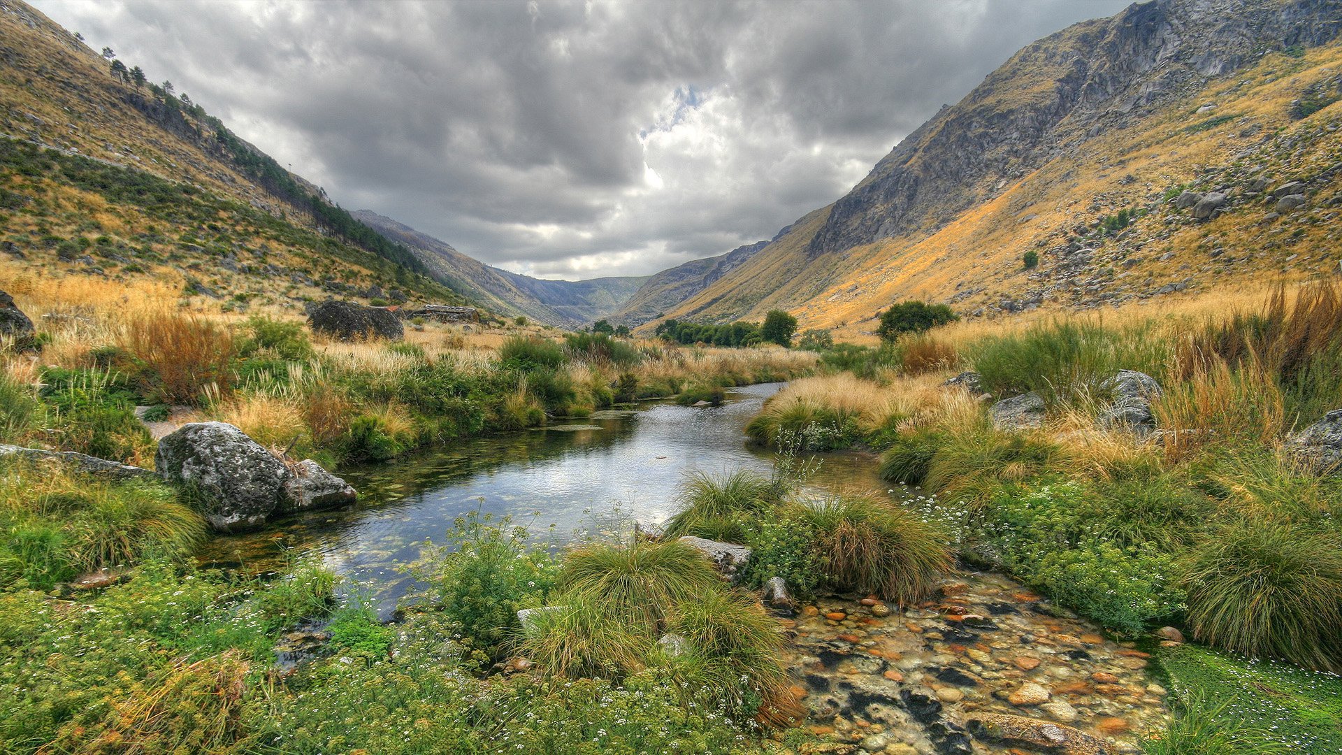 HD desktop wallpaper featuring a serene nature scene with a calm stream running through a lush green valley, flanked by rocky mountains under a cloudy sky.