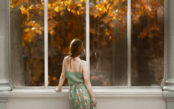 A redhead woman stands by a large window, gazing outside at autumn foliage, captured in a serene moment. This 4K Ultra HD image serves as an elegant desktop wallpaper.