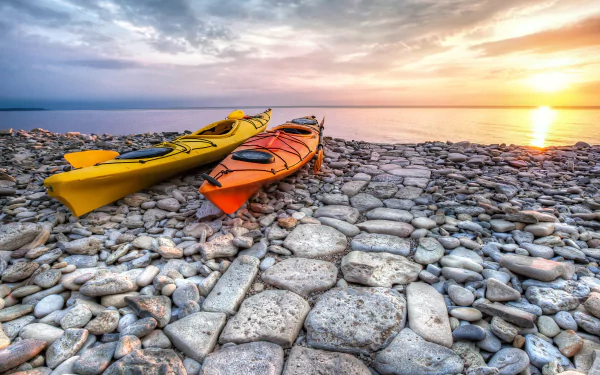 4K Ultra HD PC desktop wallpaper: two kayak vehicles resting on a stone shore at sunset, serene horizon and a glowing, pastel sky.